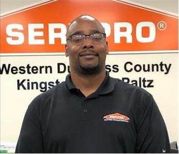 A photo of a man with glasses wearing a black polo shirt standing against a SERVPRO logo
